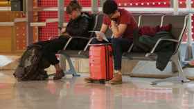 Dos personas con su equipaje esperan su vuelo sentados en un banco en el Aeropuerto de Madrid, propiedad de Aena /EP