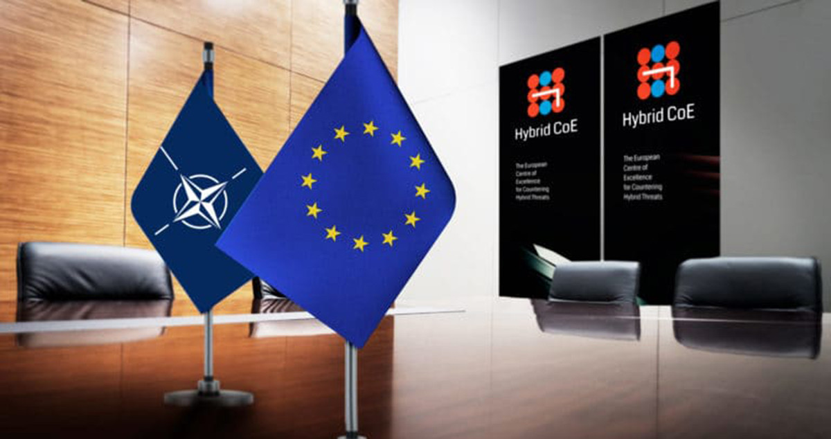 Las banderas de la UE y la OTAN ante el logo del Centro Europeo de Excelencia para Contrarrestar las Amenazas Híbridas