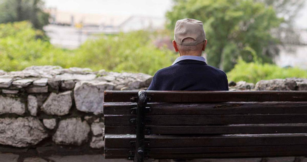Un hombre mayor en soledad sentado en un banco frente a un parque / CG