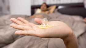Una mano aguanta un preservativo mientras un joven espera en la cama para tener sexo / PIXABAY