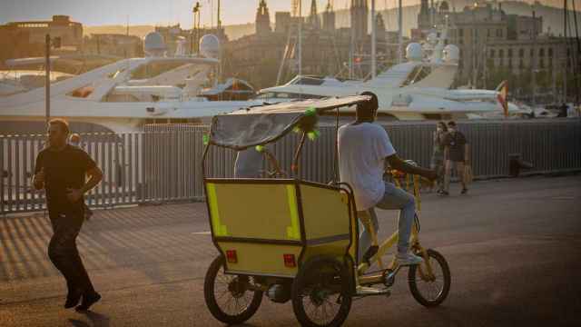 Un bici-taxi para turistas pasa por delante de los yates del Puerto de Barcelona / EP