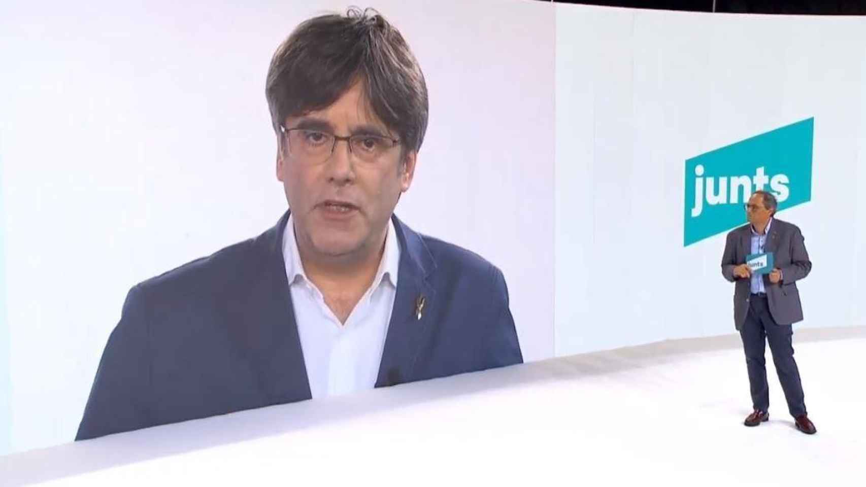 Carles Puigdemont, en pantalla, con el presidente Quim Torra, en el acto de JxCat / EP