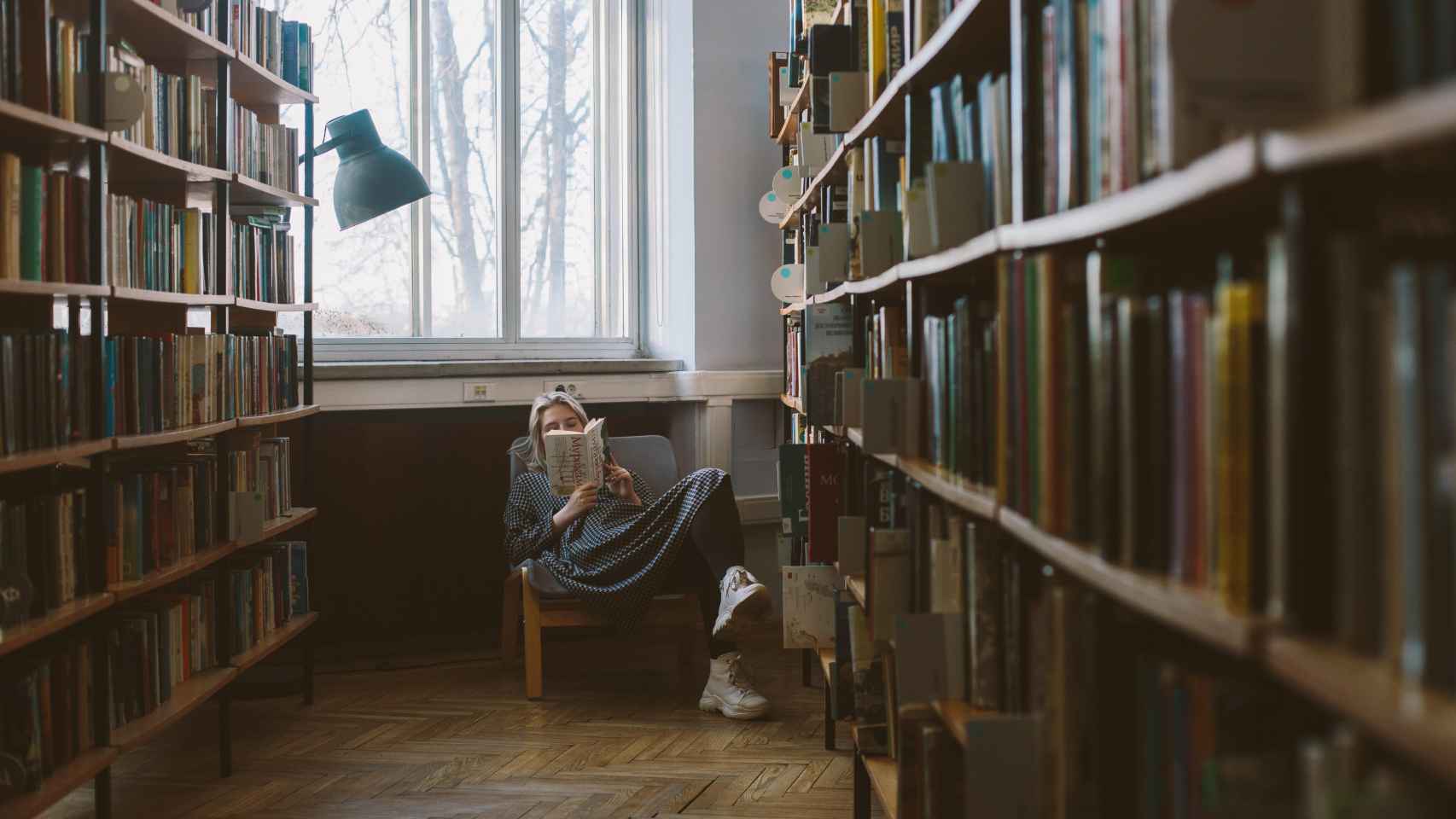 Una mujer sentada lee un libro en una biblioteca / PEXELS