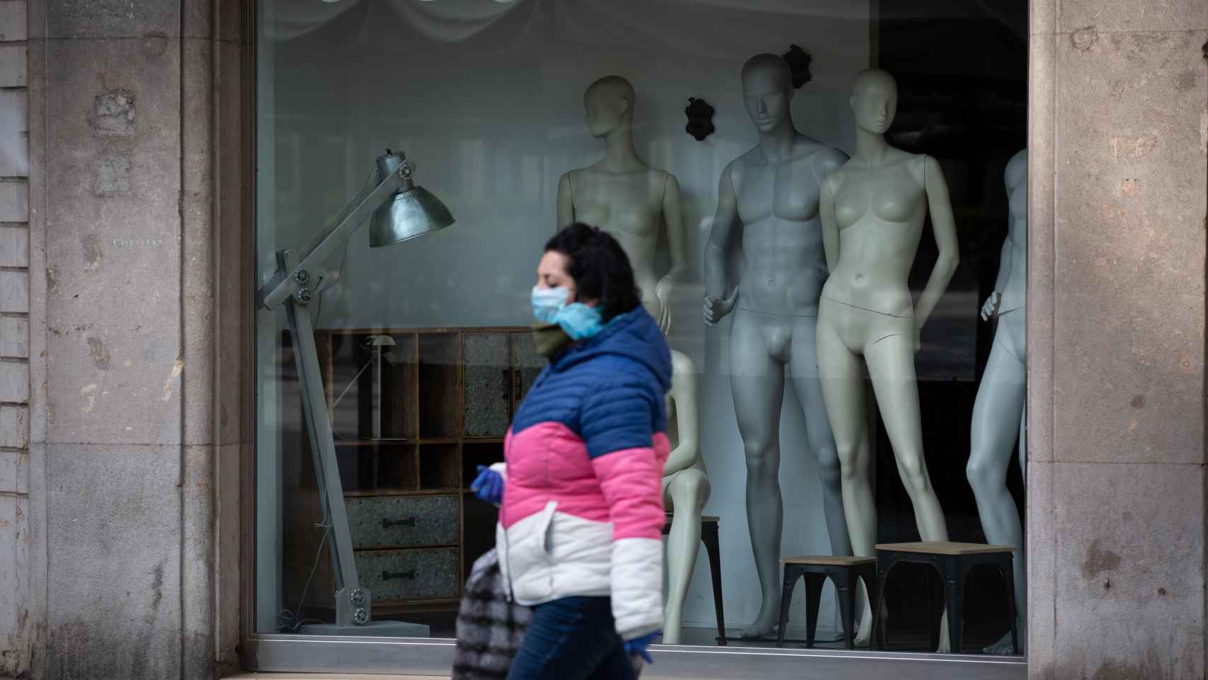 Una mujer protegida con una mascarilla pasa junto al escaparate de una tienda cerrada en Barcelona / EUROPA PRESS