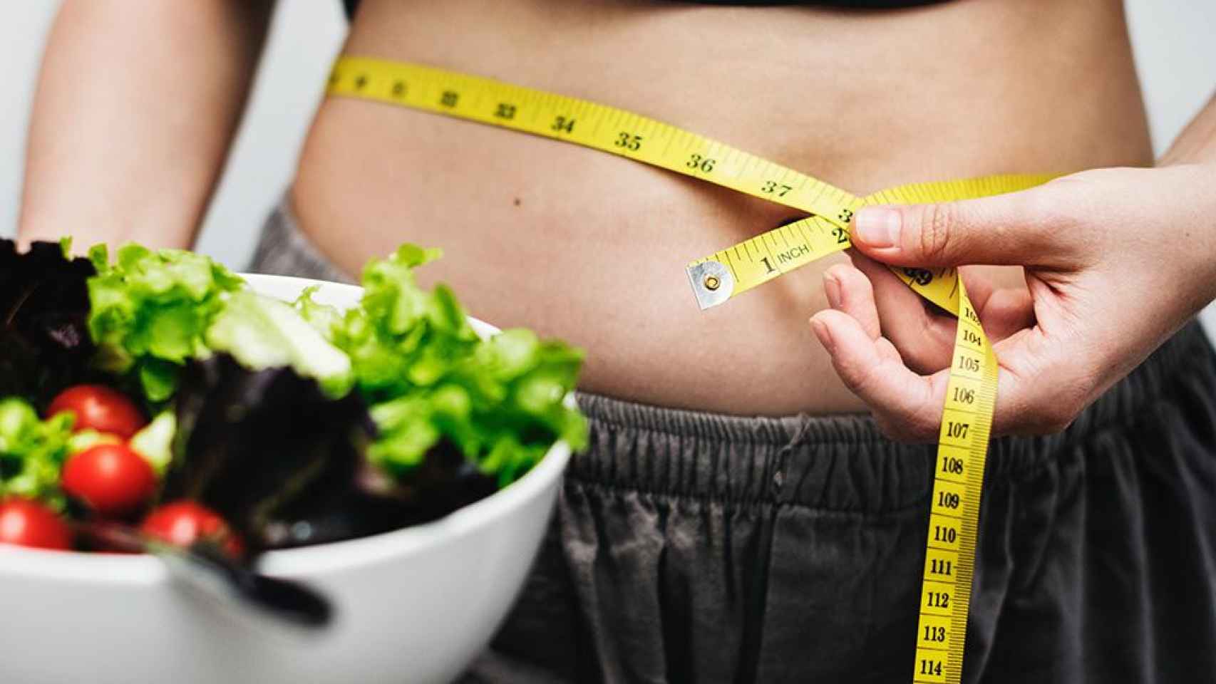 Una persona mide su cintura mientras sostiene una ensalada en una mano, que ha de combinar con complementos dietéticos