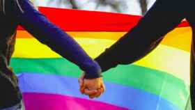 Una pareja de lesbianas cogida de la mano frente a la bandera del arcoíris