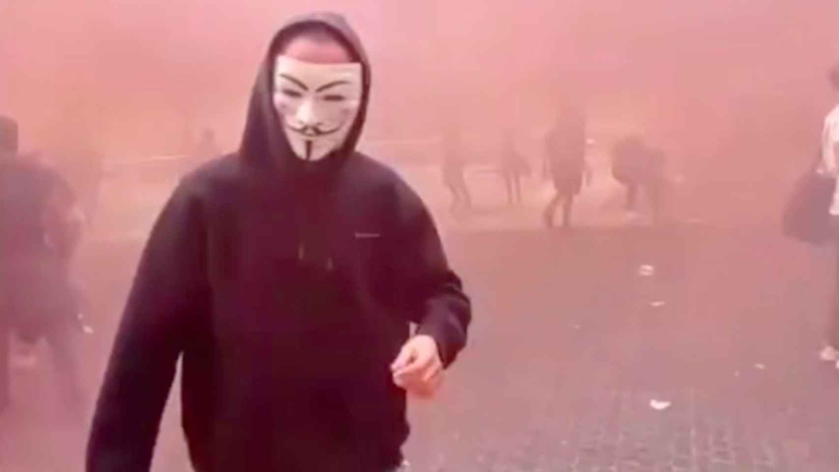 Captura de pantalla del vídeo en el que se ve a manifestantes con máscaras de Anonymus activando bombas de humo rojas y lanzando piedras en la plaza de Sant Jaume