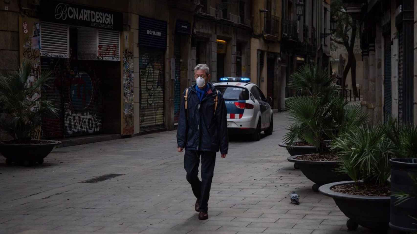 Un hombre con mascarilla pasea por una calle de Barcelona junto a un coche de la Policía / EUROPA PRESS